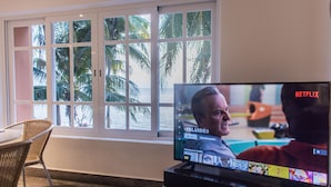 Un salon avec un téléviseur affichant l’interface de Netflix, de grandes fenêtres donnant sur des palmiers et des meubles en osier.