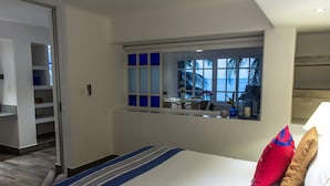 Une chambre d’hôtel avec un grand lit, une fenêtre donnant sur des palmiers, une petite table avec un vase et une armoire avec des étagères.