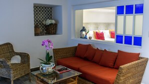 Un salon avec un canapé en osier, une table basse en verre et un fauteuil en osier. Il y a une fenêtre avec des vitres teintées en bleu.