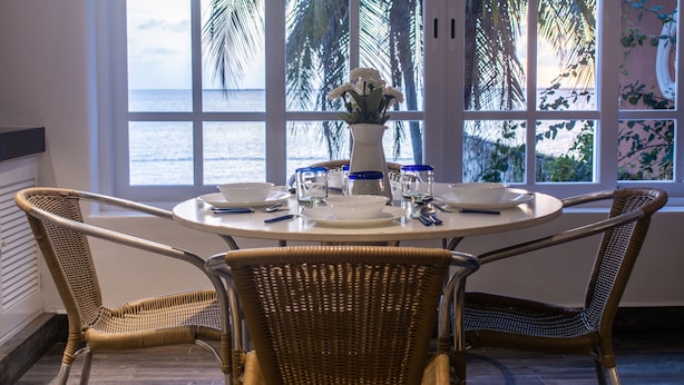 Un coin repas avec une table ronde, quatre chaises en osier, un vase rempli de fleurs et une vue sur la mer grâce à de grandes fenêtres.