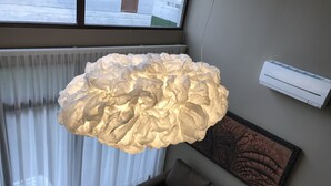 Ein Zimmer mit einer großen, strukturierten Pendelleuchte, einem Fenster mit Jalousien, einem braunen Sofa und einer Klimaanlage an der Wand.