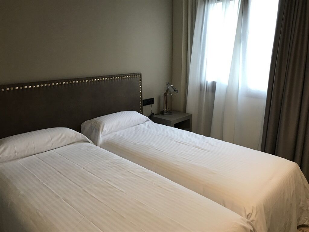 A hotel room with two single beds, a nightstand with a lamp, and a window with curtains.