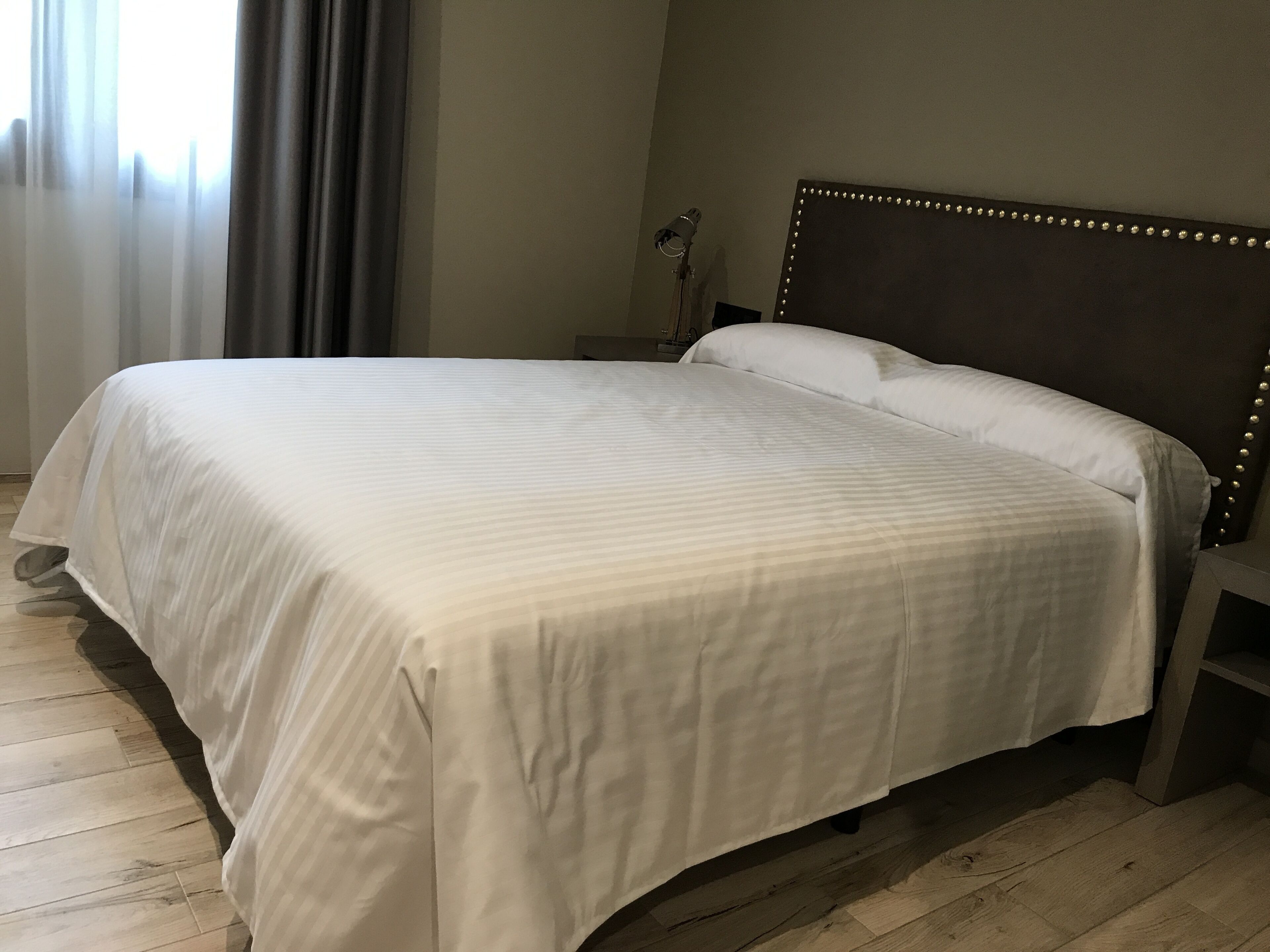 A neatly made bed with a white bedspread in a room with wooden flooring and a dark headboard with light fixtures.