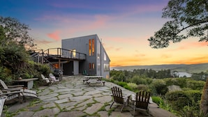 A modern house with a stone patio, outdoor seating, and a scenic view of hills and a lake at sunset. - A Sanctuary to Relax and Refresh all your Senses (Inverness)