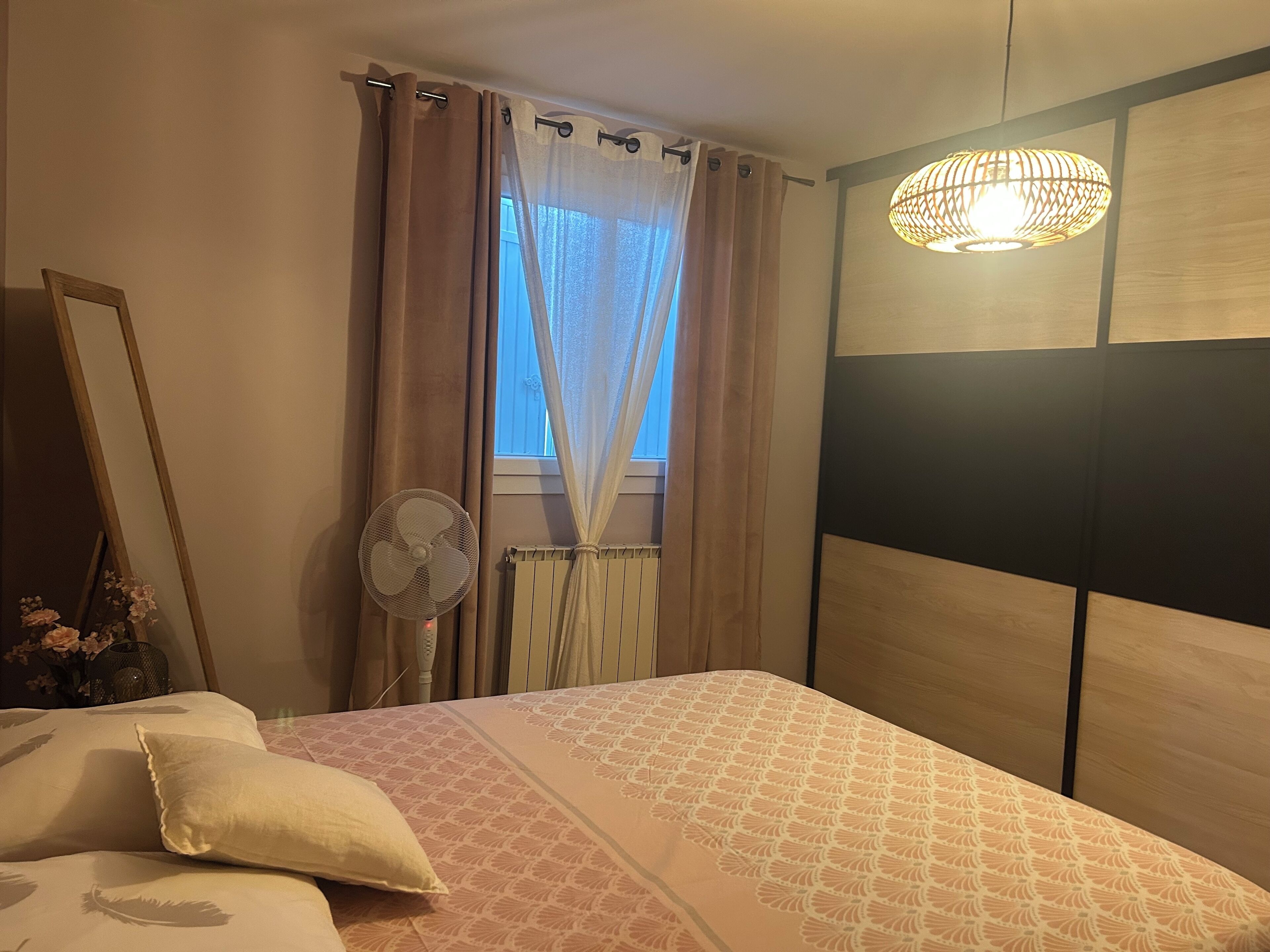 A bedroom with a bed, a bedside fan, a window with curtains, and a wall-mounted light fixture.