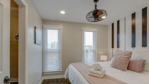 Une chambre avec un grand lit, deux fenêtres équipées de stores, un ventilateur de plafond et une porte donnant sur un couloir.