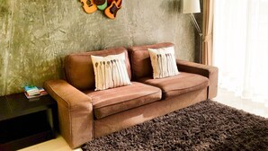 A living room with a brown sofa, two decorative pillows, a dark rug, and a wall with mounted string instruments. - Super-clean Penthouse (WIFI) - Luxury Pad with everything for the vacation (Koh Samui)