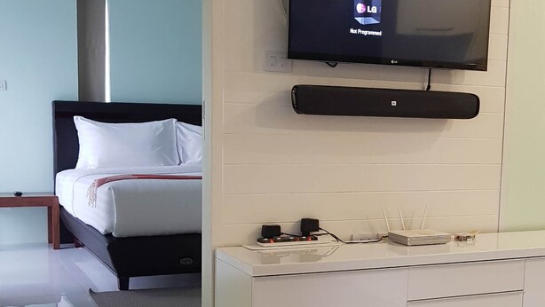 A modern hotel room with a flat-screen TV mounted on the wall, a white console table, a bed with white bedding, and a dark rug on the floor. - Super-clean Penthouse (WIFI) - Luxury Pad with everything for the vacation (Koh Samui)