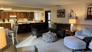 A spacious living area with a sofa, ottoman, and a dining table with chairs. A kitchen is visible in the background.