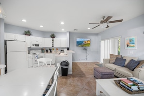 A modern kitchen with white cabinetry, a dining area with a white table, and a living room with a beige sofa and a TV mounted on the wall.
