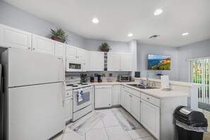 Cocina moderna con armarios blancos, horno empotrado, microondas y una isla grande con fregadero. Hay plantas en macetas sobre los armarios y un cuadro en la pared.
