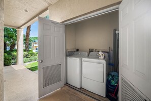 A laundry room with two washing machines and a dryer, a door leading outside, and a trash bin. - PGA Village/Castle Pines Best Location Pet Friendly. (Port St. Lucie)