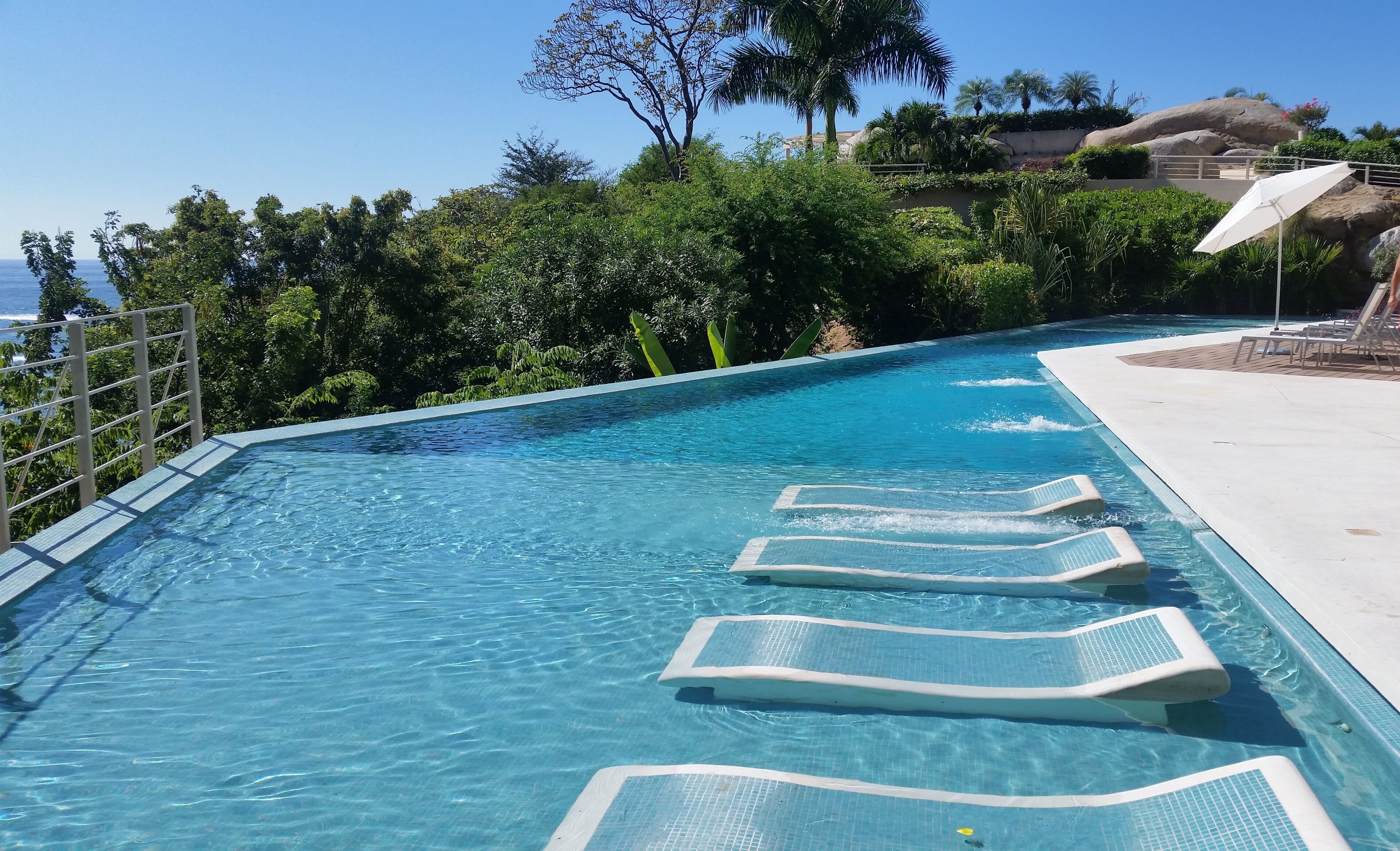 A swimming pool with clear blue water, surrounded by lush greenery and a view of the ocean.