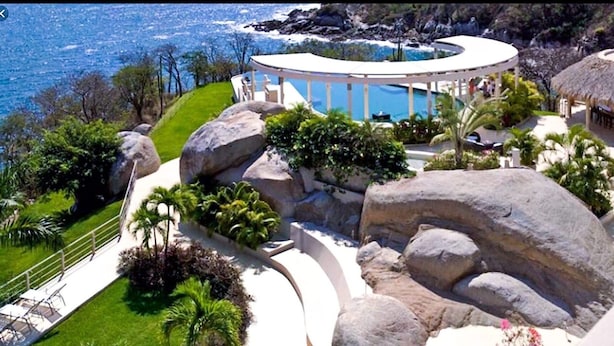 Un complexe hôtelier doté d’une grande piscine, entourée d’une végétation tropicale luxuriante et de gros rochers.