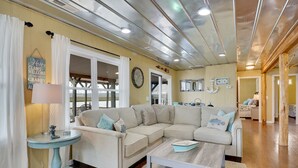 Una sala de estar con un sofá seccional color beige, una mesa redonda y una mesa de centro. Hay una ventana con cortinas blancas, un reloj de pared y un letrero que dice 'Happy Clams Welcome'.