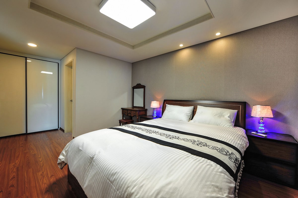 Una habitación de hotel con una cama grande, suelo de madera, una mesita de noche con lámpara, un espejo y un armario.