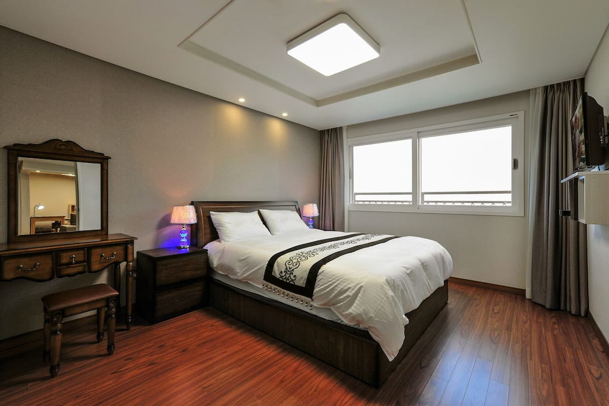 Una habitación de hotel con una cama grande, una mesita de noche con lámpara, un banco de madera, un aparador y un ventanal con cortinas.