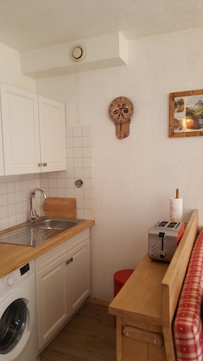 Une cuisine compacte avec des meubles blancs, un plan de travail en bois, un lave-linge, un évier, un grille-pain et une horloge murale.