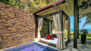 Un espace extérieur privé avec un jacuzzi, un mur en pierre servant de décor, un coin salon couvert avec des coussins et une vue sur l’océan.