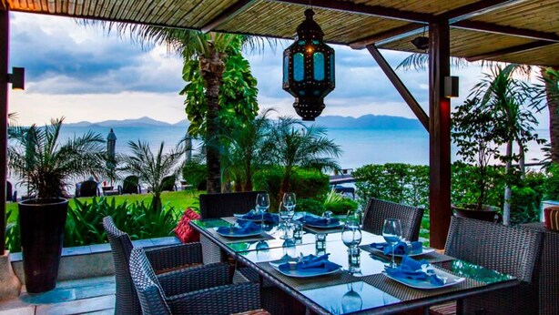 Un espace de restauration en plein air avec une table dressée pour deux personnes, entouré de chaises en osier, sous un toit de chaume avec une lanterne suspendue au-dessus.