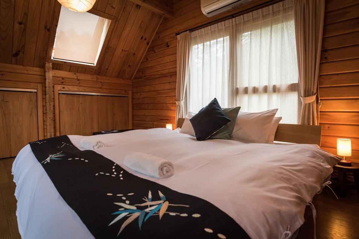 A wooden cabin bedroom with a large bed, a skylight, and a nightstand with a lamp.
