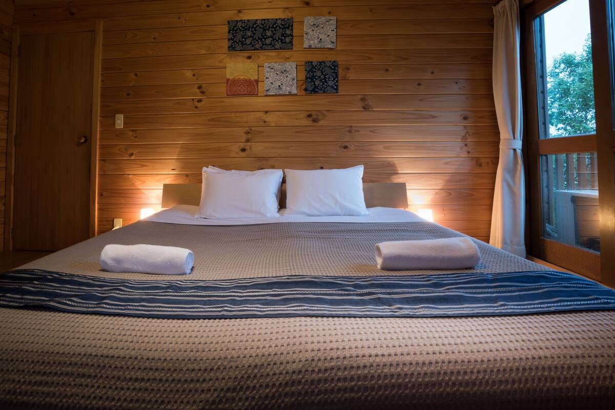A wooden-paneled room with a bed, two pillows, a blanket, and a window with curtains.
