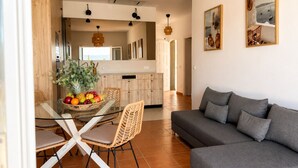A modern living area with a grey sofa, wicker chairs, a glass dining table with fruit, and a kitchen in the background.