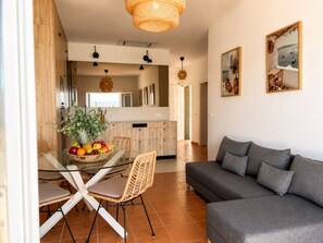 Un moderno salón con un sofá gris, sillas de mimbre, una mesa de comedor de cristal con fruta y una cocina al fondo.