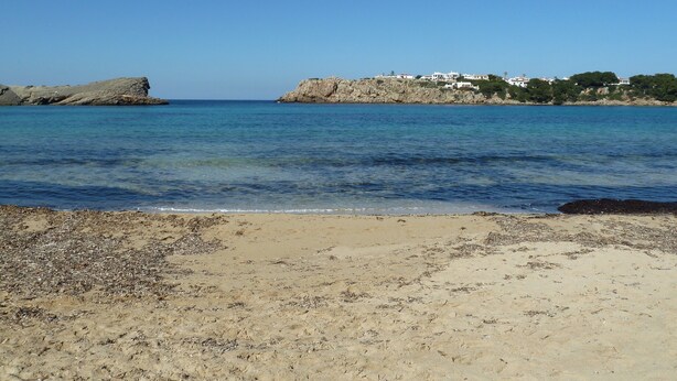 A beach with clear water, rocky outcrops, and buildings on a hillside.