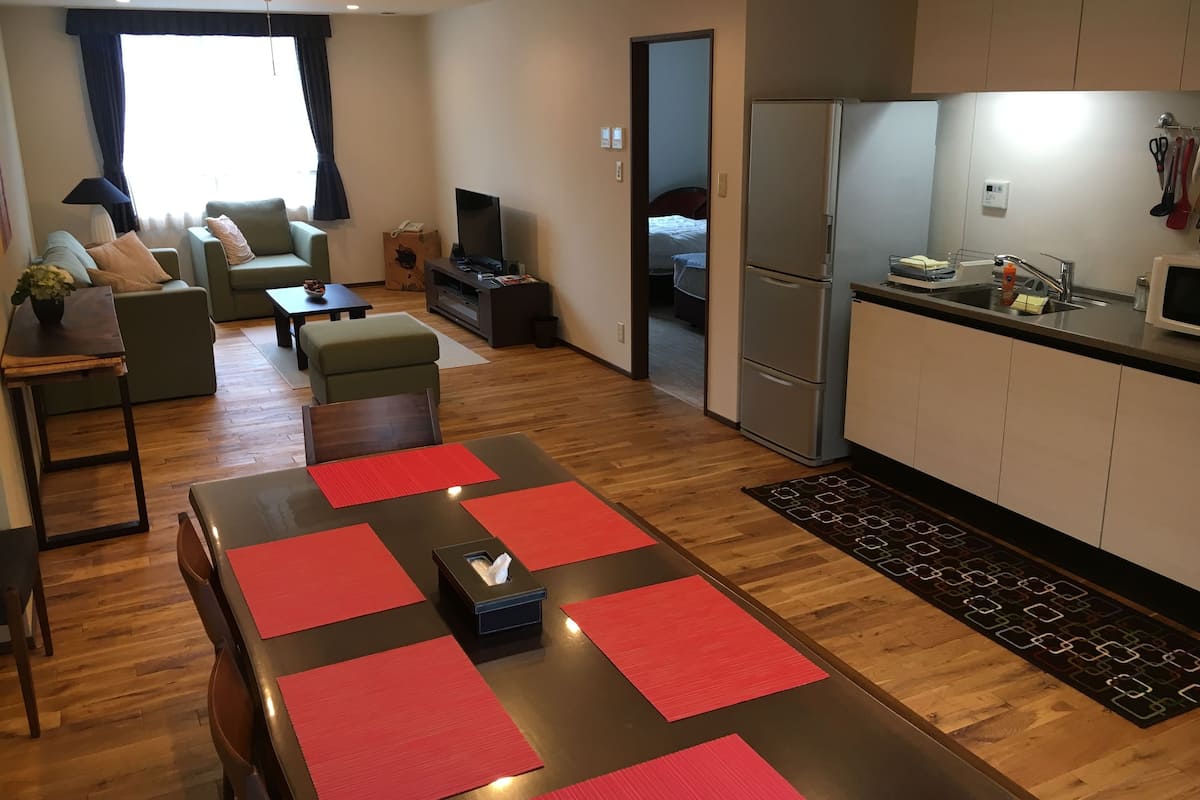 A modern living room with a sofa, a coffee table, and a TV. There is a kitchen area with a refrigerator and a sink.