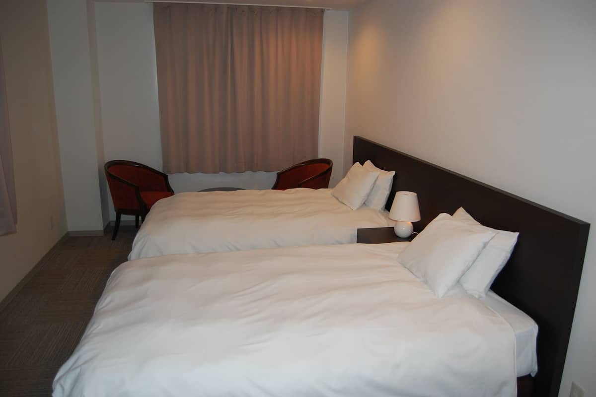A hotel room with two beds, a chair, a lamp, and a window with curtains.