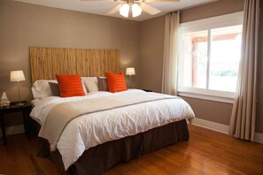 Een netjes opgemaakt bed met witte beddengoed en oranje kussens, een houten hoofdbord, een nachtkastje met een lamp, en een raam met gordijnen.