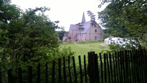 A church with a tall steeple, surrounded by trees and a fenced area with tables and chairs. - House in Breton village between land and sea (Le Cloitre Saint Thegonnec)