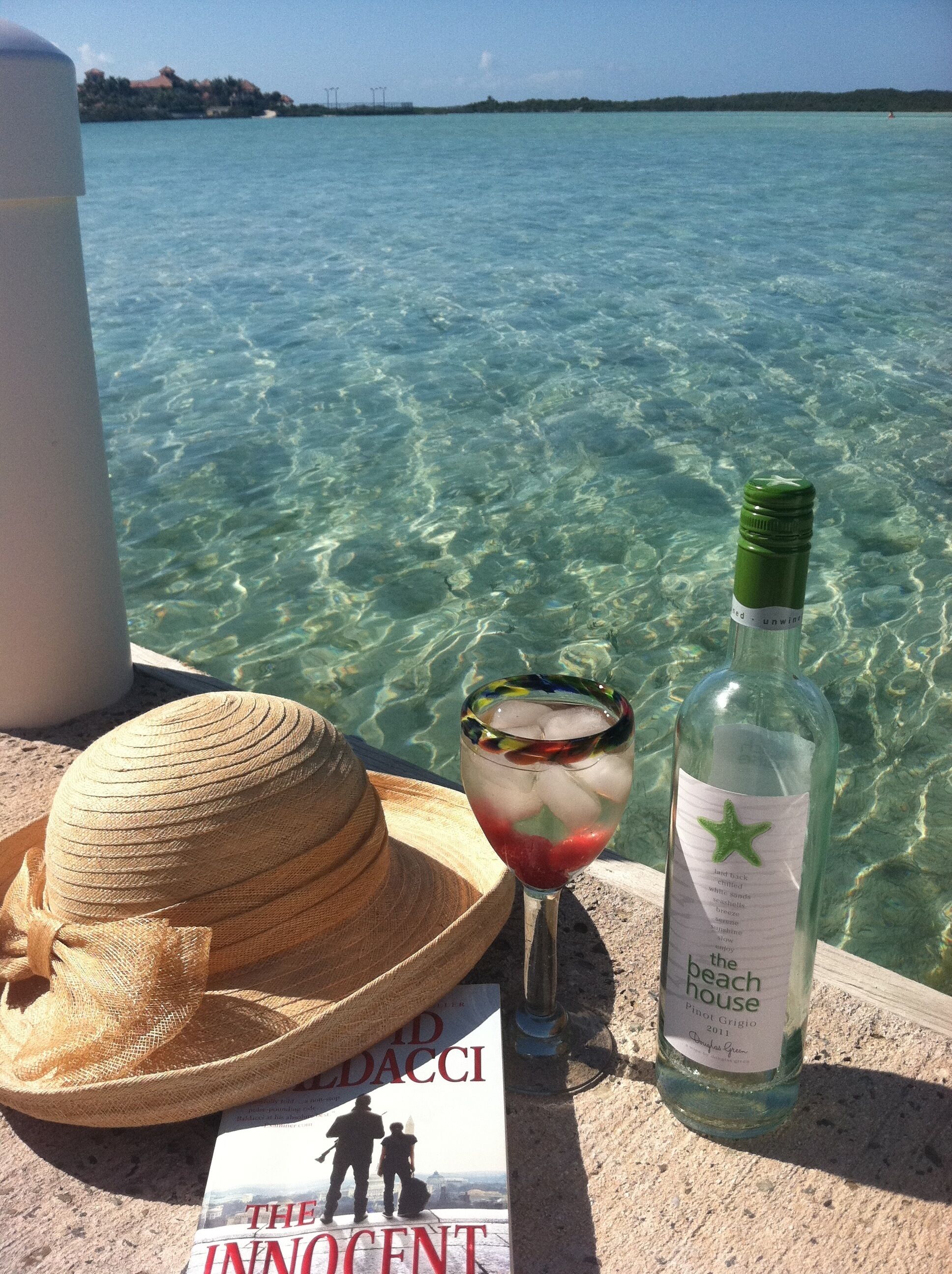 A straw hat, a glass with a drink, and a bottle of wine on a ledge by a clear blue sea.