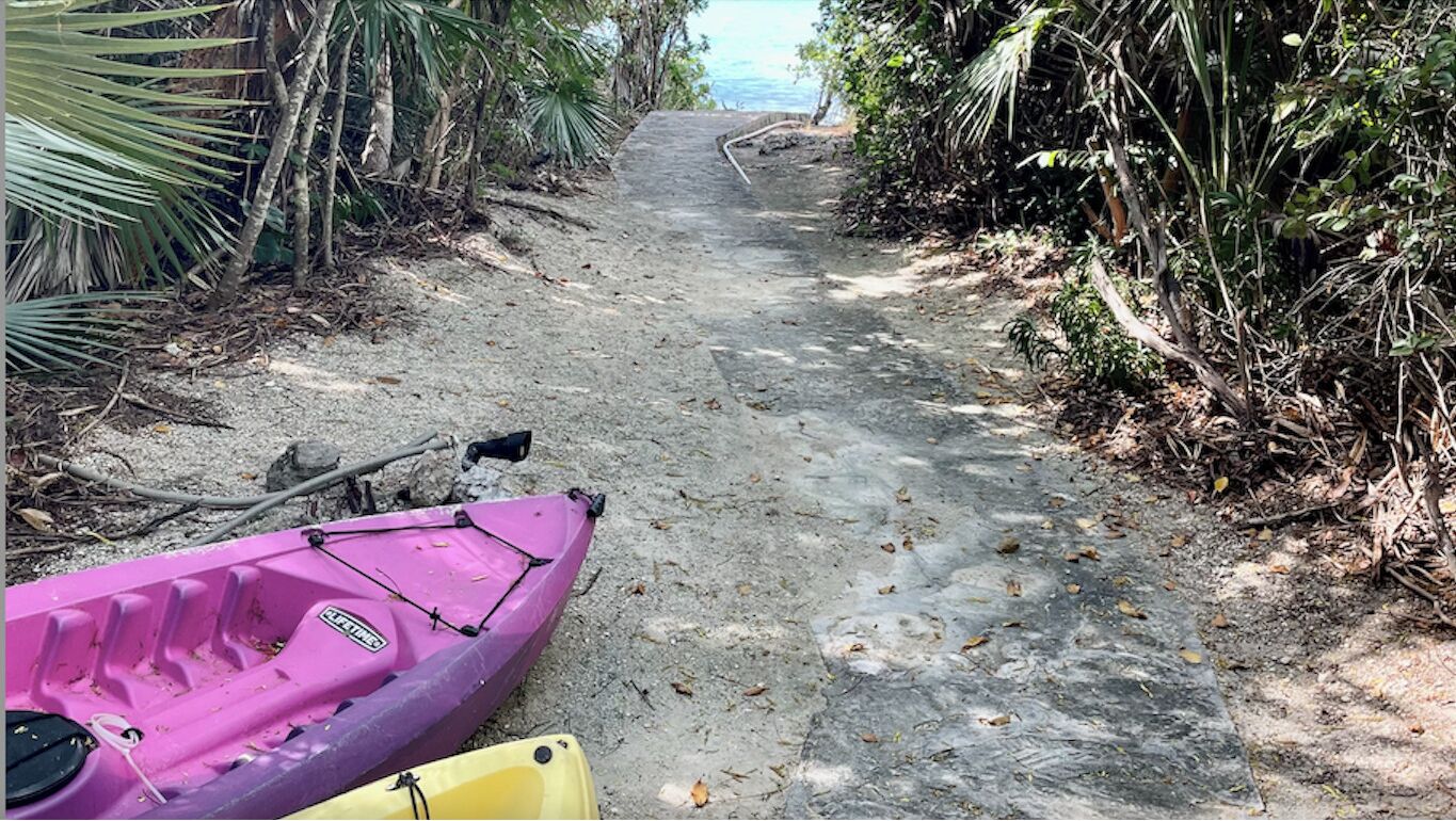 A concrete path leading to a beach, surrounded by dense vegetation. A purple kayak is parked on the left side of the path.