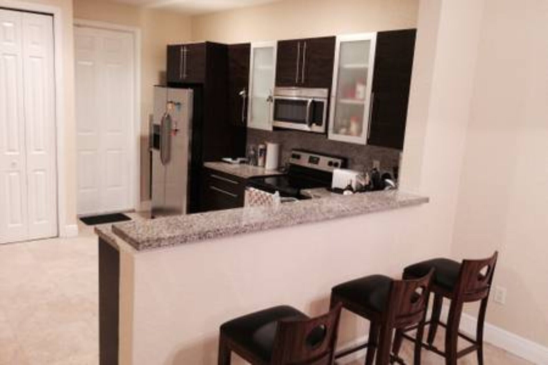 A compact kitchen with dark cabinetry, a stainless steel refrigerator, a microwave, and a stove. There are two bar stools at the counter.