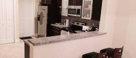 A compact kitchen with dark cabinetry, a stainless steel refrigerator, a microwave, and a stove. There are two bar stools at the counter.