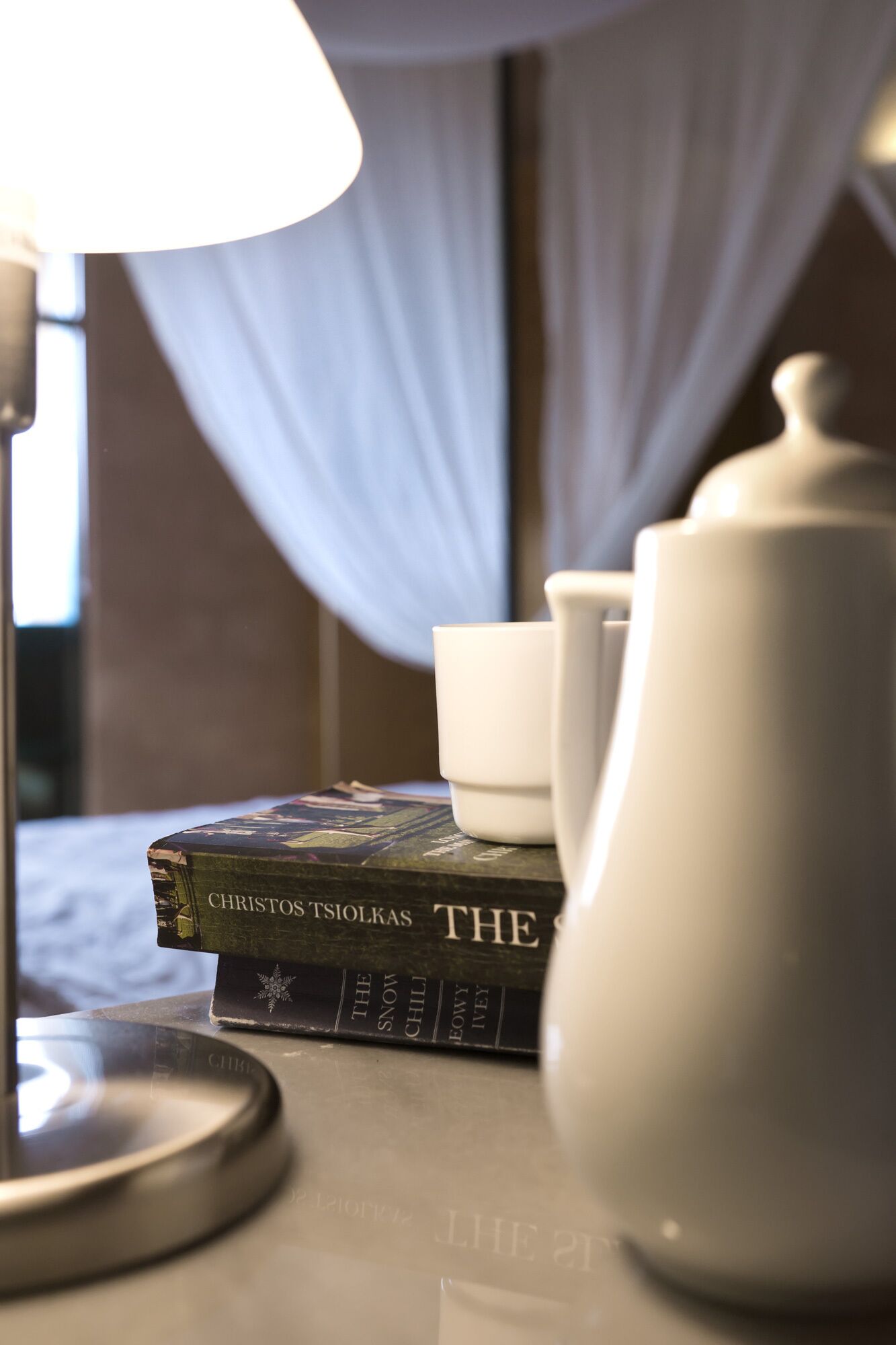 A bedside table with a lamp, a teapot, a cup, and a book titled "The Snow Child" by Christos Tsiolkas.