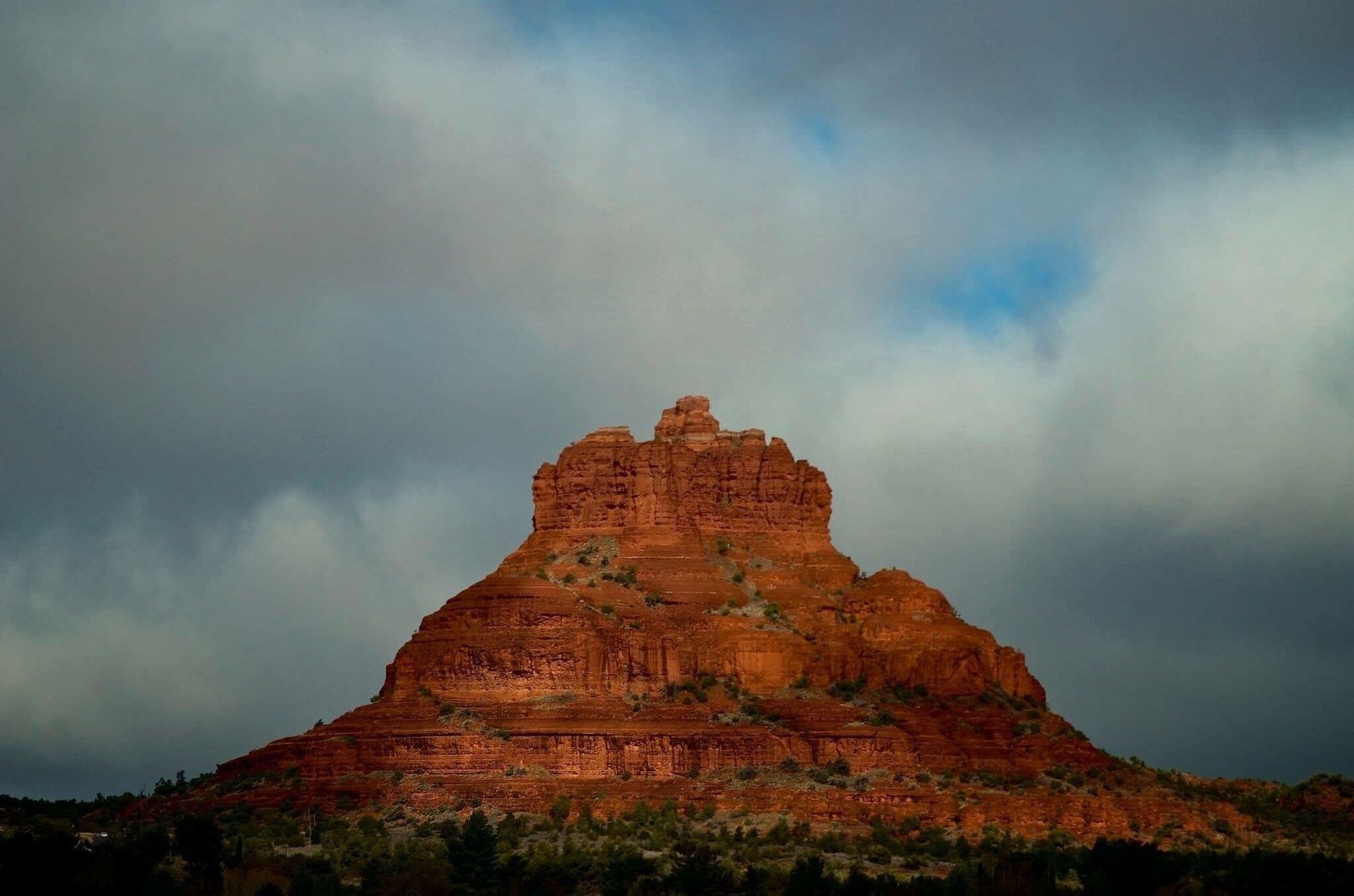 A large, red rock formation with a flat top and steep sides, surrounded by sparse vegetation under a cloudy sky.
