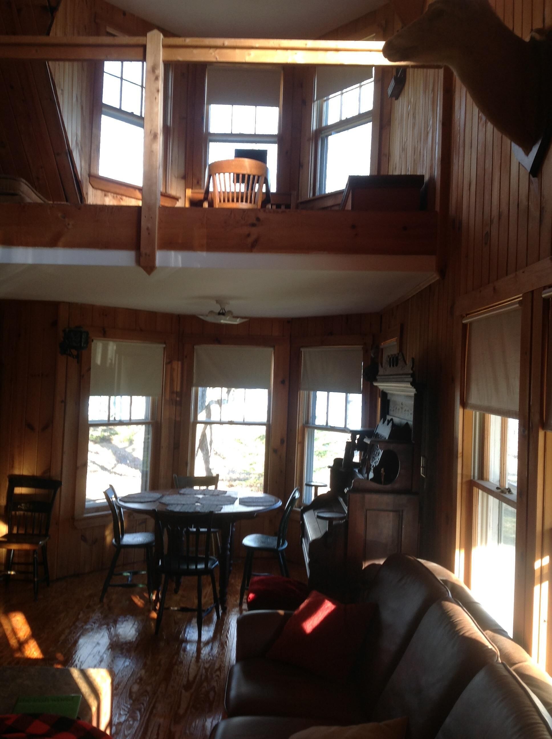 Interior de una cabaña con comedor, un altillo con una silla y zona de cocina.