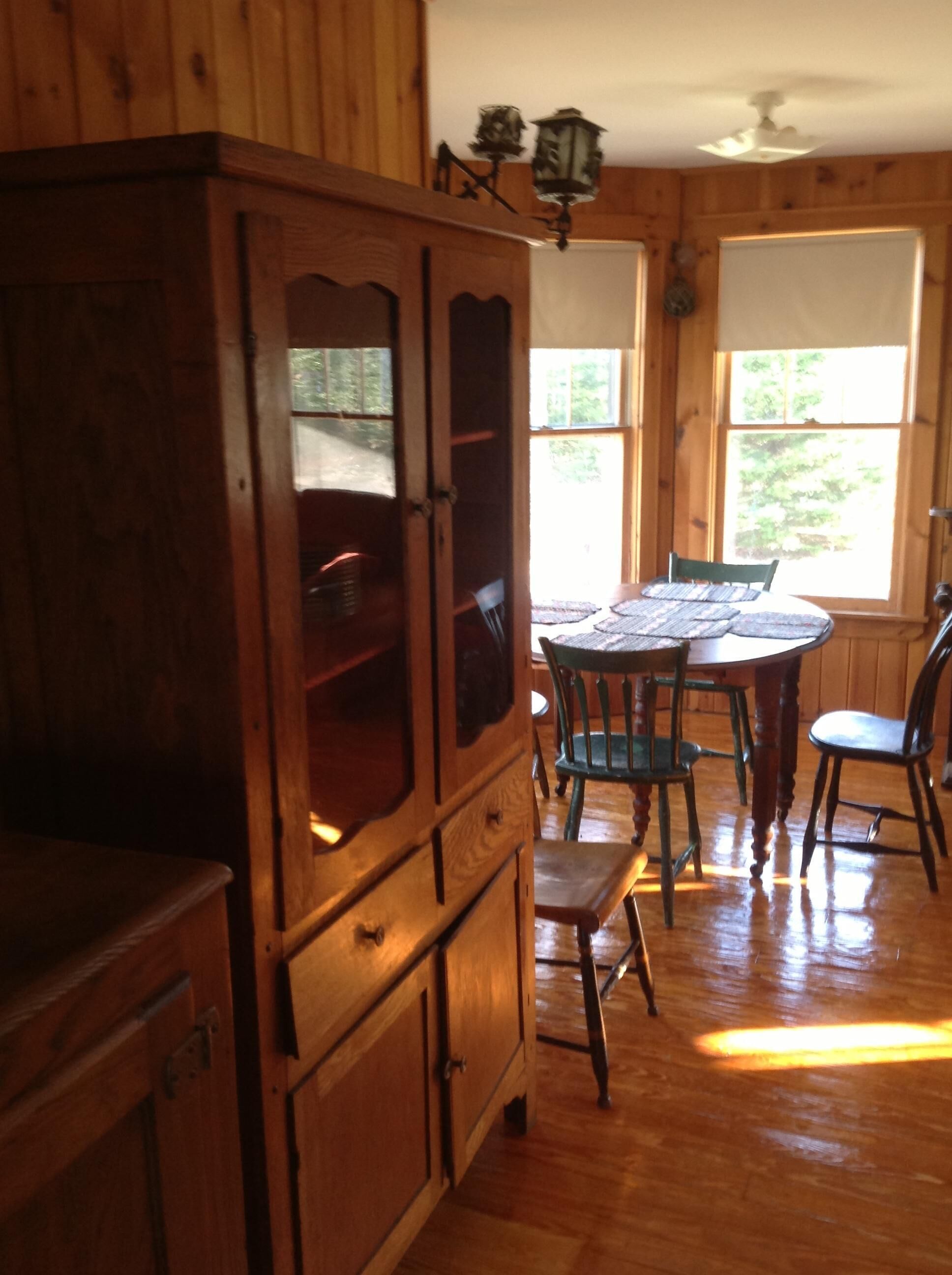 Interior de una cabaña de madera con una mesa y sillas para comer, un armario con puertas de vidrio y una ventana con vistas a follaje.