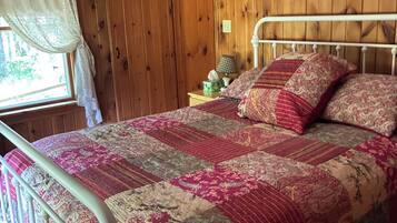 Una habitación con paneles de madera, una cama con estructura de metal, una mesita de noche con lámpara y una ventana con cortina.