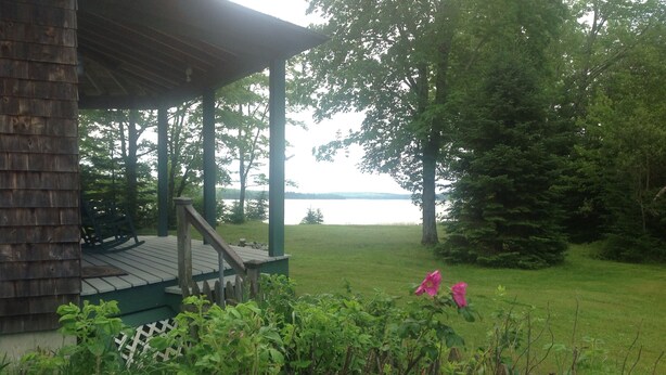 A wooden deck with a railing, overlooking a grassy area with a lake and trees in the background.