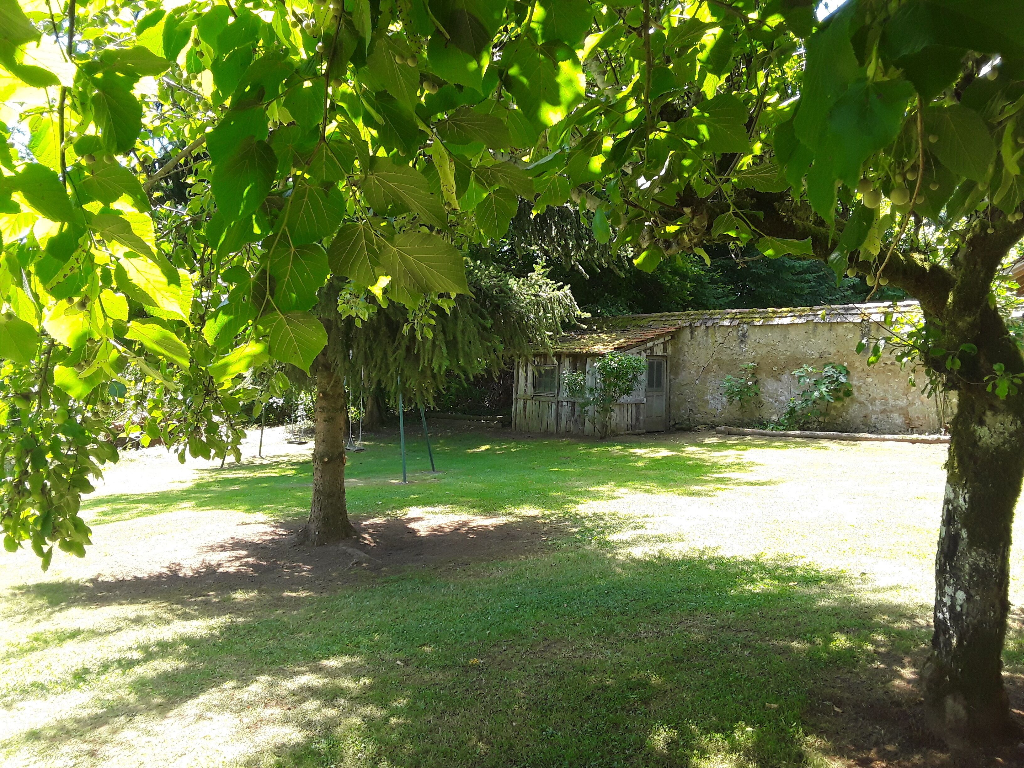 A rustic building with a stone wall and a wooden door, surrounded by a grassy yard and trees.