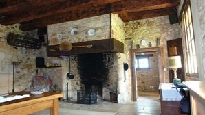 Una habitación rústica con pared de piedra, vigas de madera, chimenea, una mesa de madera y una ventana pequeña.
