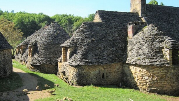Una hilera de edificios de piedra con techos de paja, un sendero cubierto de hierba y un rebaño de ovejas.
