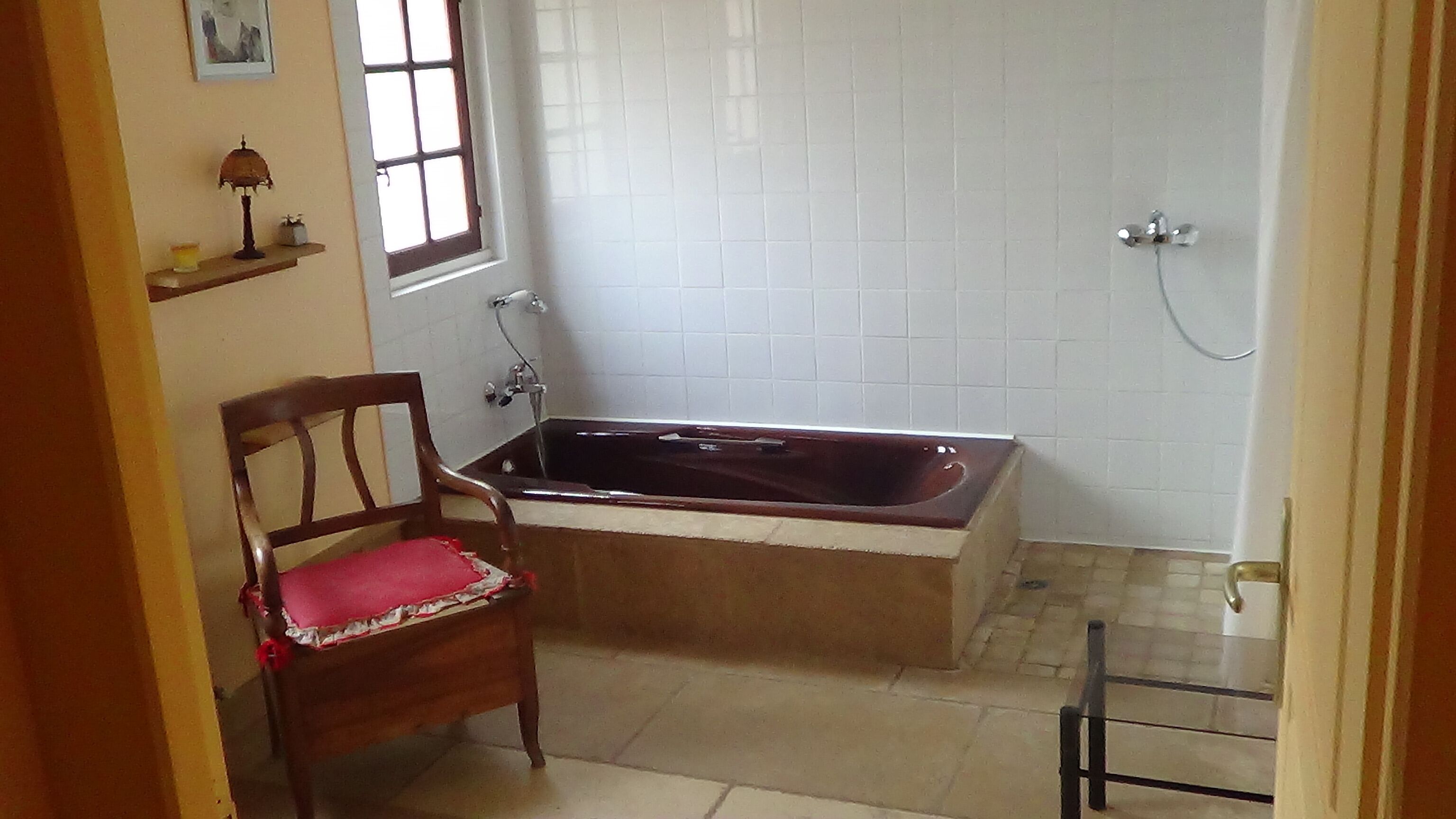 Un bagno con una vasca, una sedia di legno con cuscino rosso, una finestra e un muro rivestito di piastrelle.