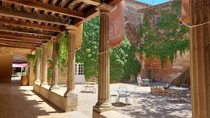 Un patio al aire libre con columnas de piedra, una pérgola y un muro de ladrillo cubierto de hiedra. Hay mesas y sillas al aire libre, y un letrero con texto.