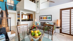 A spacious living area with a glass dining table, a sofa set, and a kitchen in the background. - Maui beachfront, steps to the sand!*Hono Kai C1* (Wailuku)
