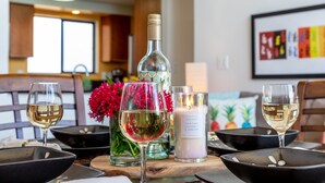 A dining table with wine glasses, a bottle, and candles in a well-lit room with wooden furniture and a window. - Maui beachfront, steps to the sand!*Hono Kai C1* (Wailuku)